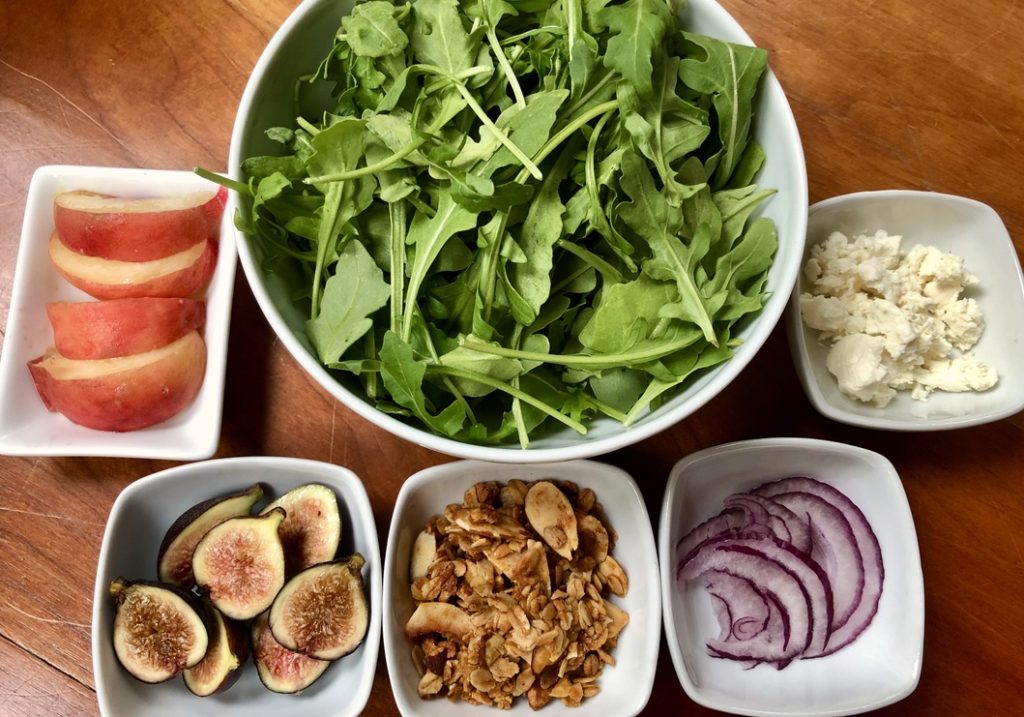 ingredients for salad:  arugula, peach slices, sliced figs, goat cheese crumbles, red onion slices, and granola